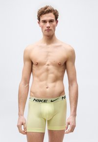 Νεαρός άνδρας με κατσαρά μαλλιά που φοράει ανοιχτό κίτρινο μπόξερ της Nike, στέκεται μπροστά σε απλό ανοιχτό φόντο.