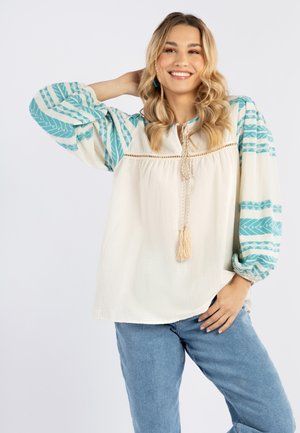 Blusa bianca con maniche a fantasia turchese, dettagli a frangia sul colletto e vestibilità rilassata. Indossata con jeans azzurri.