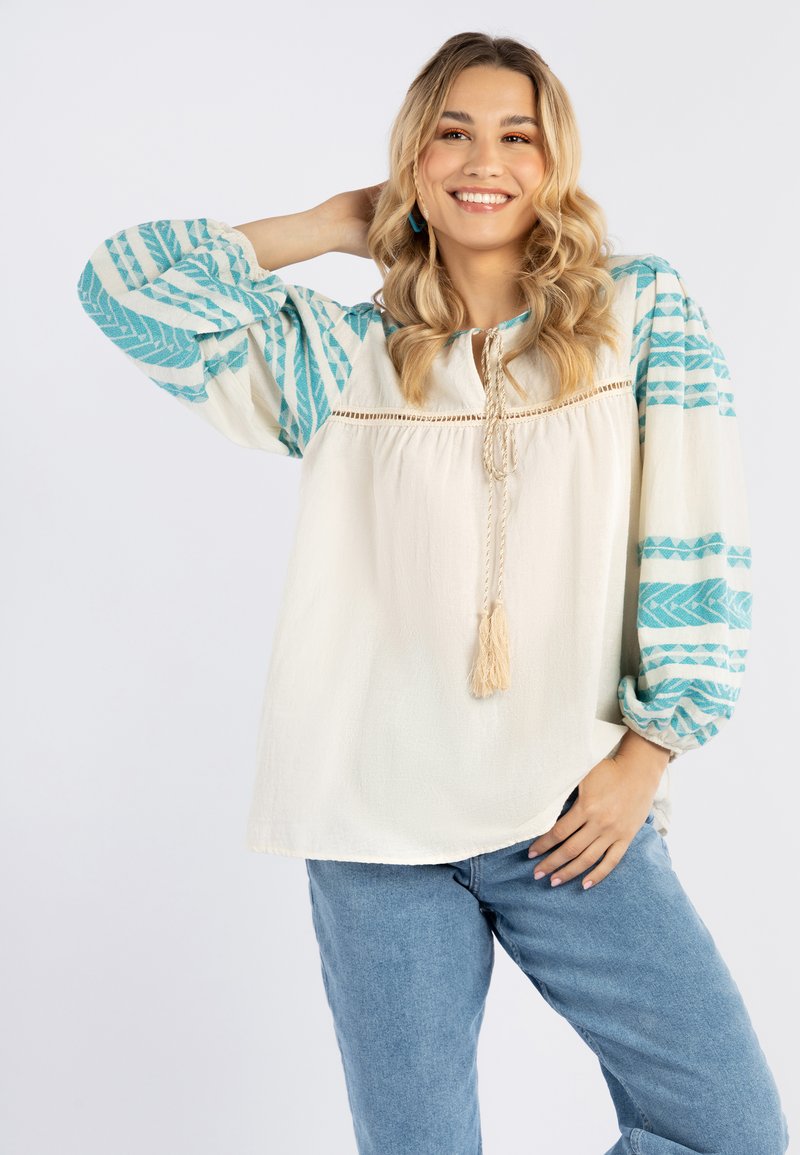 Blusa bianca con maniche a fantasia turchese, dettagli a frangia sul colletto e vestibilità rilassata. Indossata con jeans azzurri.