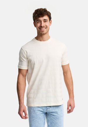 Junger Mann lächelt, trägt ein strukturiertes cremefarbenes Kurzarmhemd und hellblaue Jeans, steht vor einem schlichten weißen Hintergrund.