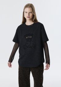 Zwarte oversized t-shirt met een tekstuur van een beer en "BOLD" in goud. Beschikt over mesh mouwen en een relaxte pasvorm. Gecombineerd met een panterprint broek.