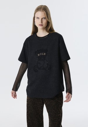 Czarny, oversize t-shirt z teksturowanym graficznym motywem niedźwiedzia i napisem "BOLD" w kolorze złotym. Posiada siateczkowe rękawy i luźny krój. W połączeniu z spodniami w panterkę.