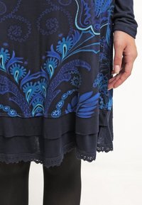 Gros plan d'une main à côté d'un ourlet de robe à motifs floraux en couches bleu marine et bleu, bordé de dentelle, portée sur des leggings noirs.