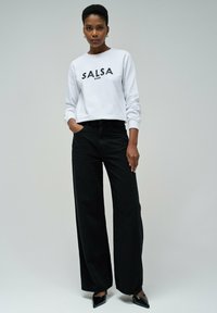 Sudadera blanca con "SALSA" en lentejuelas negras, combinada con pantalones de pierna ancha de cintura alta en negro y tacones de punta negros. Diseño simple y moderno.