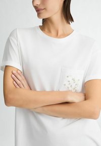 Biała bawełniana koszulka z krótkimi, wywiniętymi rękawami. Posiada kieszeń ozdobioną koralikowym wzorem. Prosty okrągły dekolt, standardowy krój.