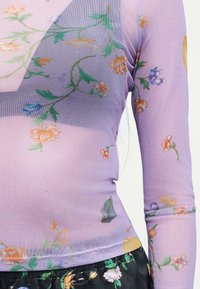 Διαφανές μακρυμάνικο τοπ λεβάντας με πολύχρωμο λουλουδένιο σχέδιο, φορεμένο πάνω από ένα πιο σκούρο εσώρουχο με αντίστοιχο ενδύμα με λουλουδένιο μοτίβο κάτω από αυτό.