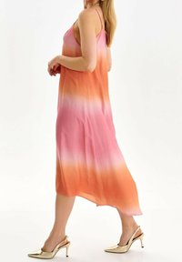 Lange, mouwloze jurk in gradient tinten van roze naar oranje, met een vloeiende vormgeving en zachte stof. Zilveren hoge hakken sandalen.