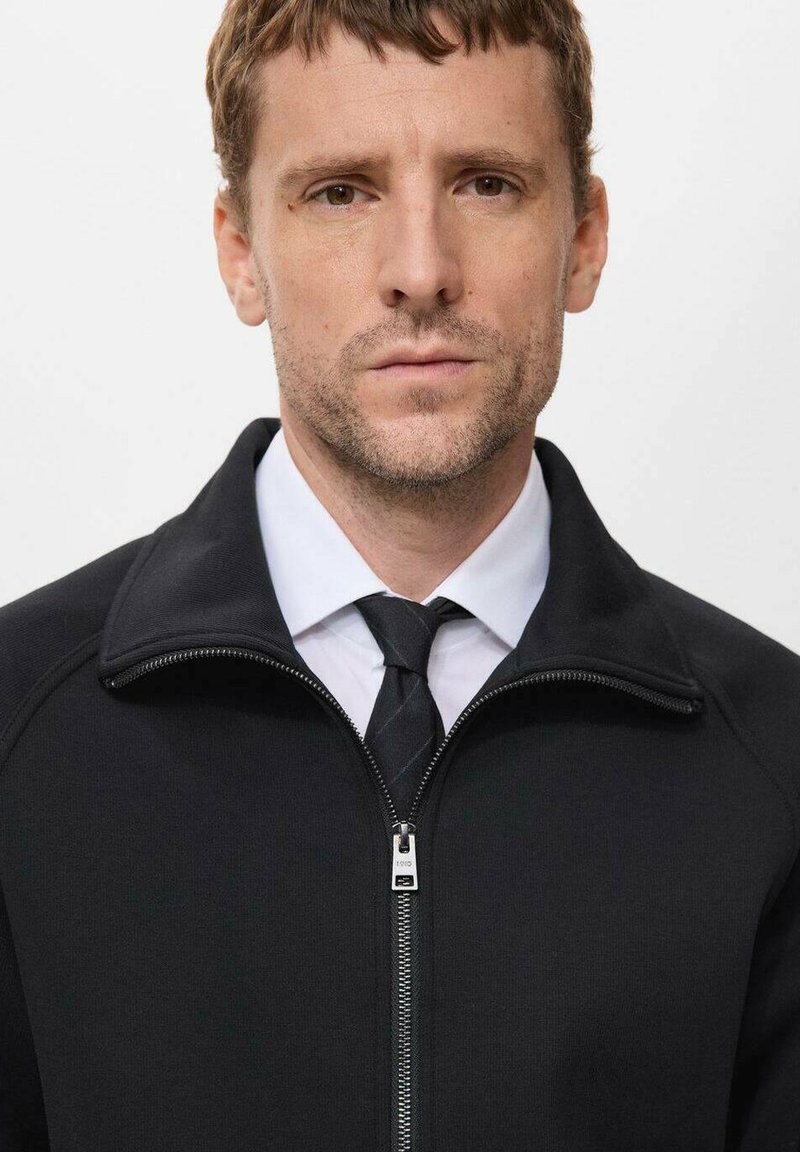 Schwarze Zip-Up-Jacke mit hohem Kragen, kombiniert mit einem weißen Hemd und einer schwarzen Krawatte. Verfügt über einen metallischen Reißverschluss und gerippte Textur.