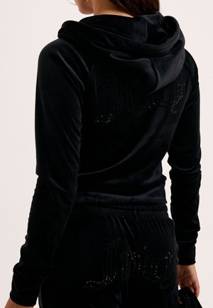 Frau trägt schwarzen Velours-Hoodie und Hose mit rhinsteinbesetzten "Juicy"-Logos auf dem Rücken, hält ein passendes schwarzes Accessoire in der rechten Hand.