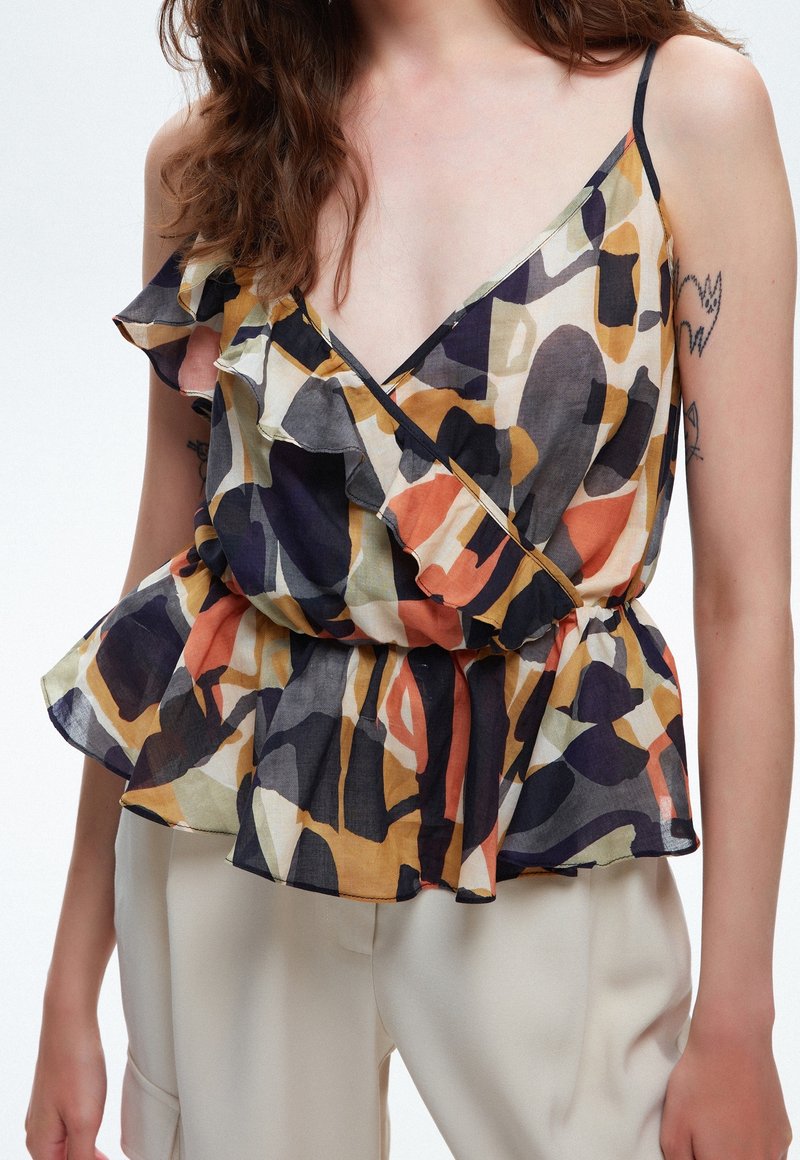 Femme portant un chemisier sans manches à motif abstrait avec des volants et un pantalon beige, montrant des tatouages sur les bras et des cheveux bruns ondulés.