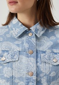 Chaqueta de mezclilla azul claro con un patrón floral, cierre con botones, cuello puntiagudo y dos bolsillos en el pecho con detalles metálicos.