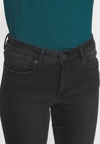 Jeans de mezclilla negra con un corte ajustado, que cuentan con cinco bolsillos, un cierre de botón negro y una textura suave. Combinados con una blusa color verde azulado.