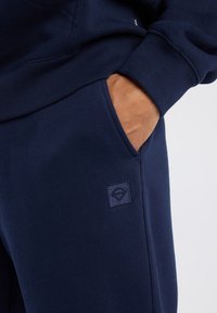 Pantalones de chándal azul marino con una textura suave, que cuentan con un bolsillo lateral y un pequeño parche de logo bordado en la parte frontal.