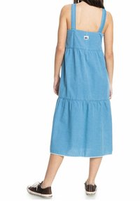 Lichtblauwe denimjurk met brede bandjes, gelaagde ontwerp, knielengte zoom en een klein logolabel aan de achterkant. Gedragen met donkere sneakers.