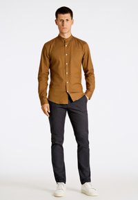 Camisa marrón de botones con cuello mandarín, diseño ajustado, mangas largas y botones blancos, combinada con pantalones oscuros y zapatillas blancas.
