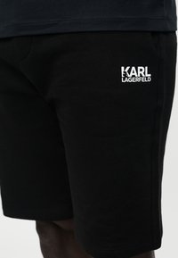 Gros plan sur un short noir avec le logo blanc "KARL LAGERFELD" sur la cuisse supérieure, porté avec une chemise noire.