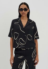 Chemise noire à manches courtes avec des motifs abstraits beiges, fabriquée en tissu texturé. Elle possède un col à cran et une fermeture à boutons à l'avant.