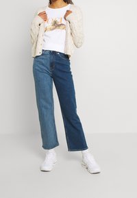 Osoba ubrana w dwukolorowe dżinsy, białą koszulkę z grafiką psa, beżowy sweter robiony na drutach, białe skarpetki i białe sneakersy stoi na jasnym tle.