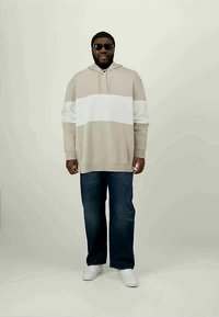 Homem com óculos de sol pretos, hoodie bege e branco, jeans azul escuro e ténis brancos, de pé contra um fundo claro e liso.
