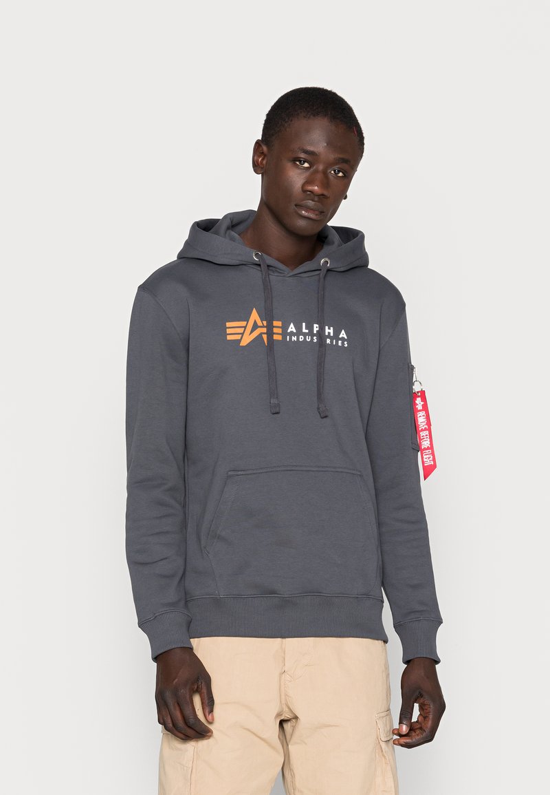 Felpa grigia con cappuccio e coulisse, tasca frontale e logo arancione di Alpha Industries. Presenta polsini e orlo a costine, include un'etichetta rossa sulla manica.