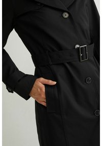 Czarny trencz z paskiem w talii. Posiada podwójnie zapięty przód, duże guziki oraz gładką teksturę. Zawiera boczne kieszenie.