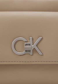 Beżowa skórzana torebka z srebrnym metalowym logo o okrągłym i kanciastym wzorze. Teksturowana powierzchnia z widocznymi detalami przeszyć.