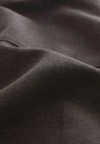 Tmavě hnědá látka zblízka, texturovaný povrch s jemným tkaninovým vzorem. Materiál působí měkce a mírně leskle, ukazuje jemné vlákna.