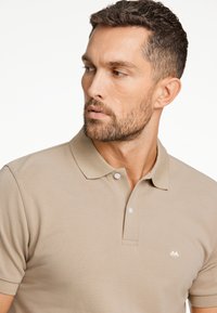 Beige polo skjorta gjord av texturerat tyg, med en klassisk krage, treknapps knäppning och en liten vit logga på bröstet.