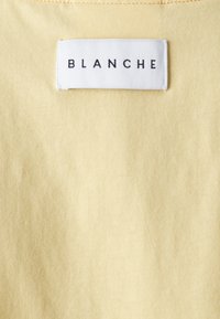 Gelbes Gewebe mit glatter Textur, das oben ein weißes Etikett aufweist, auf dem das Wort "BLANCHE" in schwarzen Buchstaben eingestickt ist.