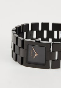 Svart klokke med firkantet urskive, gullfargede timemarkører og en kraftig lenkebånd. Elegant, matt finish med minimalistisk design.