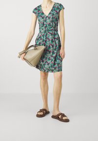 Kwiatowa zielona sukienka z dekoltem w serek, kapturem na ramionach i dopasowaną talią, zestawiona z brązowymi plecionymi sandałami i beżową torebką.