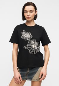 Jeune femme portant un t-shirt noir à manches courtes avec des contours floraux blancs et une jupe en jean bicolore avec une fermeture éclair à l'avant.