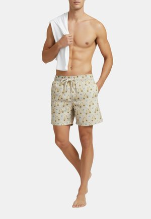 Uomo senza maglietta in piedi a piedi nudi che indossa pantaloncini da bagno beige con stampa di bottiglia gialla e cubetto di ghiaccio blu, tiene un asciugamano bianco sulla spalla.