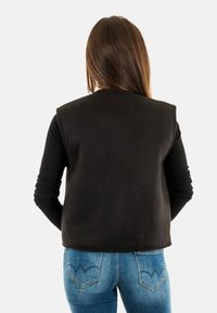 Gilet nero senza maniche con una superficie liscia, spalle arrotondate e un design pulito, abbinato a jeans blu con tasche decorative.