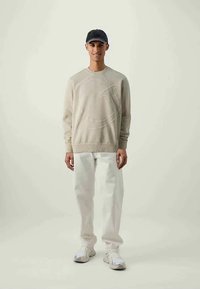 Joven con sudadera beige con un gran logotipo en relieve, pantalones blancos, zapatillas blancas y gorra negra, de pie frente a un fondo claro y simple.