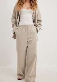 Kvinna som bär beige vida byxor och matchande jacka över en vit tanktop, står med händerna i fickorna mot en enkel bakgrund.