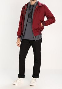 Giacca bomber bordeaux indossata sopra una t-shirt grigia, jeans neri e scarpe da ginnastica bianche. La giacca presenta una fodera interna a quadri e polsini a costine.