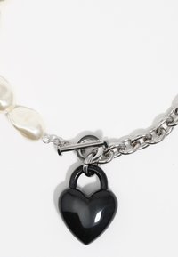 Czarny wisiorek w kształcie serca w postaci zamka na srebrnym łańcuszku, ozdobiony nieregularnymi białymi perłami oraz detalem z zapięciem typu toggle.