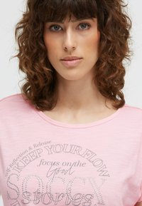 Young woman with curly brown hair wearing a pink shirt with text "KEEP YOUR FLOW" and "SORRY Stories" embellished with small beads.