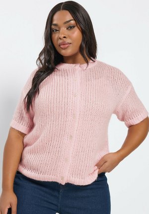 Eine Frau mit langen, dunklen Haaren trägt eine locker sitzende, kurzärmelige rosa Strickjacke und dunkelblaue Jeans und posiert mit einer Hand in der Hüfte.