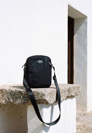 Un sac bandoulière noir Replay repose sur un rebord en pierre rugueux, à côté d’une porte encadrée de stuc blanc.