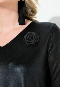 Zwarte jurk met een V-hals ontwerp, voorzien van een textuur stof en een zwarte stoffen rozenaccent aan de linkerkant, aangevuld met een lange zwarte franjes oorbel.