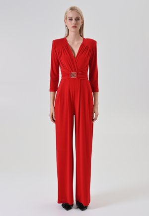 Woman stands wearing a red long-sleeve jumpsuit with a deep V-neck, belted waist, wide-leg pants, and black pointed heels on a plain background.