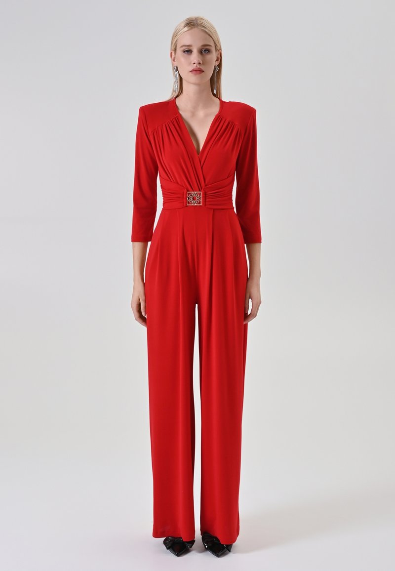 Una donna indossa una tuta rossa a maniche lunghe con scollatura a V profonda, vita cintata, pantaloni a gamba larga e tacchi a punta neri su uno sfondo semplice.