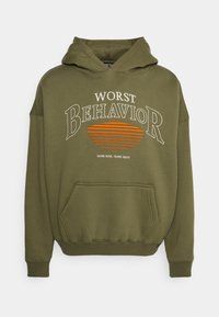 Felpa verde oliva con tasca a marsupio. La scritta in evidenza legge "WORST BEHAVIOR" in strisce bianche e arancioni. Tessuto morbido, vestibilità rilassata.