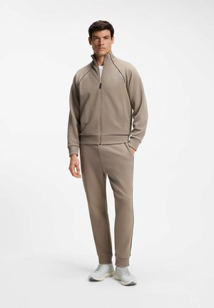 Man standing wearing beige zip-up jacket and matching pants with white sneakers on plain white background.