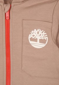 Felpa marrone con una cerniera rossa, dotata di tasca anteriore e di un logo di un albero bianco sul lato sinistro. Tessuto morbido con una superficie liscia.
