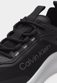 Μαύρο αθλητικό παπούτσι Calvin Klein με υφή στο επάνω μέρος, χοντρές μαύρες κορδόνια, θηλιά για κράτημα και λευκή σόλα με κυματιστό πέλμα.