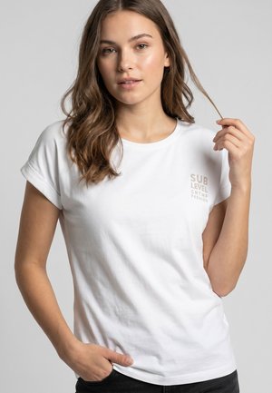 Young woman with long brown hair wearing a white T-shirt with rolled sleeves and subtle lettering on the chest, holding a hair strand.