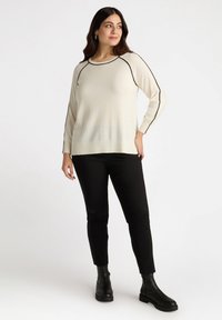 Elfenbeinfarbene Strickpullover mit schwarzem Besatz, langen Ärmeln und geripptem Saum. Getragen mit schwarzen, figurschmeichelnden Hosen und knöchelhohen Stiefeln, präsentiert von einem Modell.